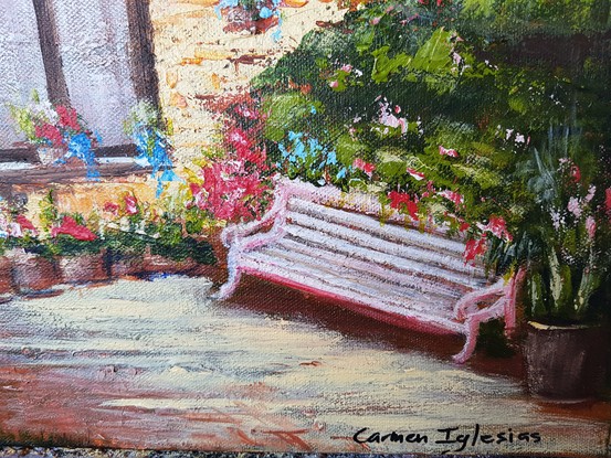 A pinkish/white park bench among colourful flowers. The bench is near an ochre colour wall on which you can see two large windows with white voile curtains and dark brown frames. Several pots with flowers hang from the wall. 