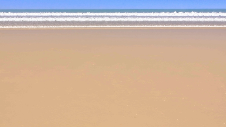 the photograph shows a smooth sandy beach on a sunny day with three waves breaking the shore