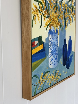 Branches of yellow wattle in a blue and white vase, with blue glass bottles and a pile of books.