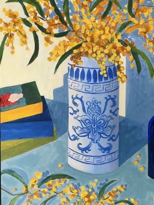 Branches of yellow wattle in a blue and white vase, with blue glass bottles and a pile of books.