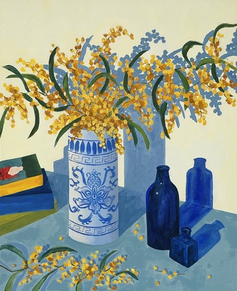 Branches of yellow wattle in a blue and white vase, with blue glass bottles and a pile of books.