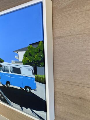 Blue Kombi van in carpark near large trees, both casting strong shadows in the foreground with hint of ocean in the background under and clear blue sky. 