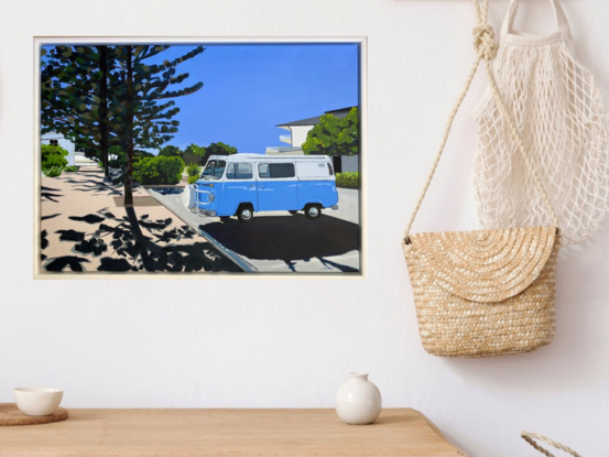 Blue Kombi van in carpark near large trees, both casting strong shadows in the foreground with hint of ocean in the background under and clear blue sky. 