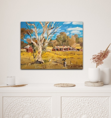 A scenic view of rural outback Australia. Includes a large white gum tree in the foreground with old sheds under a bright blue cloudy sky.