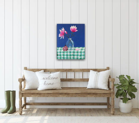 A still life depicting - a glass vase with two magnolia flowers and buds, a pomegranate leaning against the vase and a single fallen petal sitting nestled into the gingham creased table cloth. With a bold pacific blue backdrop 