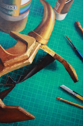 A still life looking down onto a crafting table with a green cutting mat, a scattering of paint cans and tubes, some brushes and a segmental cutting blade. The scene is dominated by a gold foam headpiece with curved horns. In the shadows is a cup of black coffee.