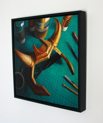 A still life looking down onto a crafting table with a green cutting mat, a scattering of paint cans and tubes, some brushes and a segmental cutting blade. The scene is dominated by a gold foam headpiece with curved horns. In the shadows is a cup of black coffee.