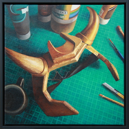 A still life looking down onto a crafting table with a green cutting mat, a scattering of paint cans and tubes, some brushes and a segmental cutting blade. The scene is dominated by a gold foam headpiece with curved horns. In the shadows is a cup of black coffee.