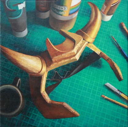 A still life looking down onto a crafting table with a green cutting mat, a scattering of paint cans and tubes, some brushes and a segmental cutting blade. The scene is dominated by a gold foam headpiece with curved horns. In the shadows is a cup of black coffee.