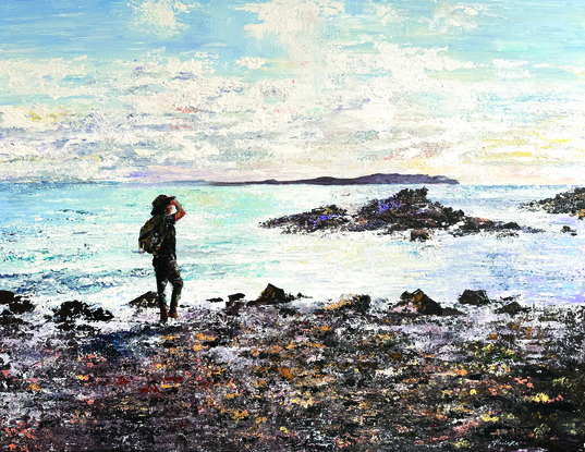 Man standing on rocky beach edge wearing a hat looking out towards a small island just offshore. 