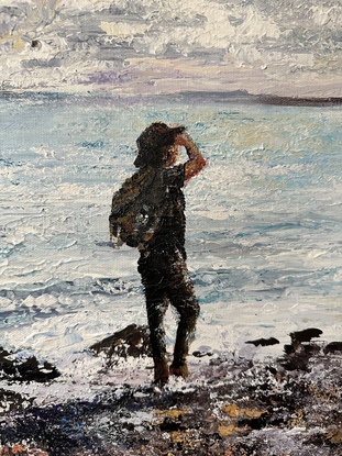 Man standing on rocky beach edge wearing a hat looking out towards a small island just offshore. 