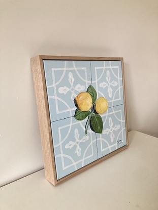 Two lemons, attached to their stems and resting on a powder blue encaustic tile background, accented by a contrasting shadow