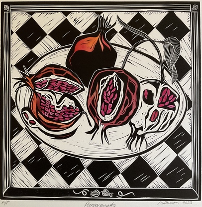 A square format black and white linoprint hand coloured in shades of red watercolour. There are four pomegranates on a plate on a black and white check tablecloth. 