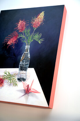 Two pinks and orange grevillia flowers in a glass bottle vase. One grevillia flower lays beside the glass bottle next to a pink paper crane. The glass bottle looks like the classic coca-cola bottle, but the text on the bottle has changed to 'Belle Australis', meaning beautiful Australia.