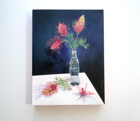 Two pinks and orange grevillia flowers in a glass bottle vase. One grevillia flower lays beside the glass bottle next to a pink paper crane. The glass bottle looks like the classic coca-cola bottle, but the text on the bottle has changed to 'Belle Australis', meaning beautiful Australia.