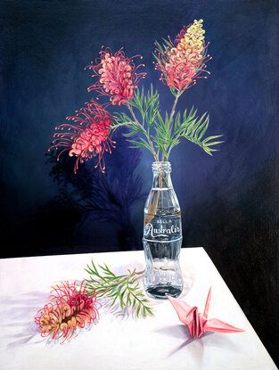 Two pinks and orange grevillia flowers in a glass bottle vase. One grevillia flower lays beside the glass bottle next to a pink paper crane. The glass bottle looks like the classic coca-cola bottle, but the text on the bottle has changed to 'Belle Australis', meaning beautiful Australia.