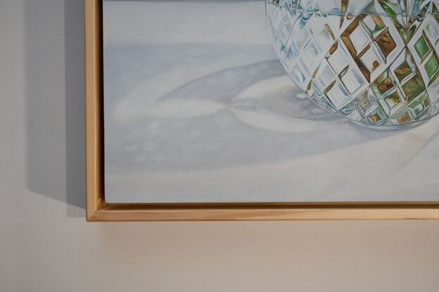 A large white magnolia flower and leaves of green and gold, in a cut crystal round bowl shaped vase. The background is a light grey white colour. The painting is framed by a light coloured wooden floating frame.