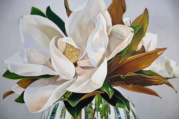 A large white magnolia flower and leaves of green and gold, in a cut crystal round bowl shaped vase. The background is a light grey white colour. The painting is framed by a light coloured wooden floating frame.