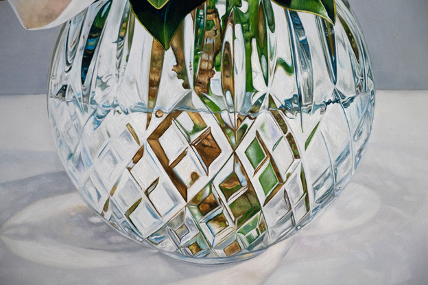 A large white magnolia flower and leaves of green and gold, in a cut crystal round bowl shaped vase. The background is a light grey white colour. The painting is framed by a light coloured wooden floating frame.