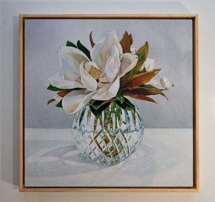 A large white magnolia flower and leaves of green and gold, in a cut crystal round bowl shaped vase. The background is a light grey white colour. The painting is framed by a light coloured wooden floating frame.