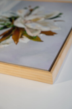 A large white magnolia flower and leaves of green and gold, in a cut crystal round bowl shaped vase. The background is a light grey white colour. The painting is framed by a light coloured wooden floating frame.