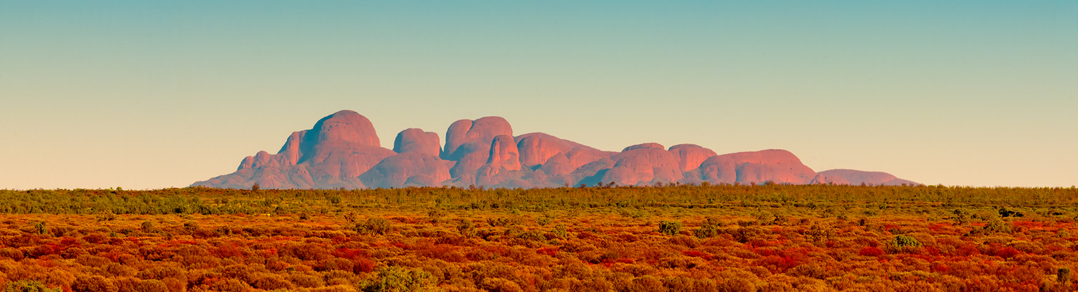 Panoramic photo of Mount Olga in Australia's Northern Territory, featuring stunning mountain range backdrop against rugged, red-hued foreground.