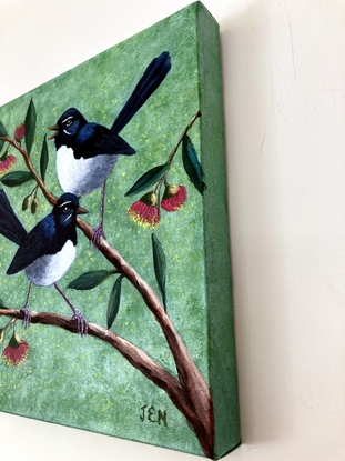 Two beautiful Willy Wagtails sitting on a eucalyptus tree with red and yellow gum blossoms. The background is a pale green speckled colour, with specks of yellow blossom sifting through the air.  The two birds appear to be in conversation.