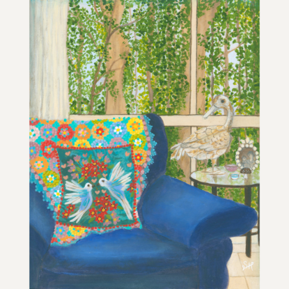 Interior scene with a rusty duck ornament on a glass side table, next to a blue arm chair and a rainbow crochet blanket hanging over the back behind a blue cushion featuring white birds and pink and red flowers. Against a window backdrop featuring leafy green trees