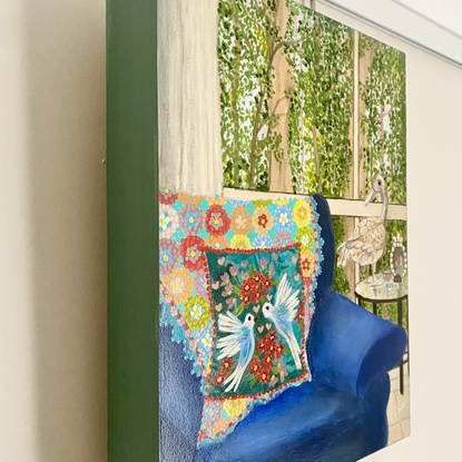 Interior scene with a rusty duck ornament on a glass side table, next to a blue arm chair and a rainbow crochet blanket hanging over the back behind a blue cushion featuring white birds and pink and red flowers. Against a window backdrop featuring leafy green trees