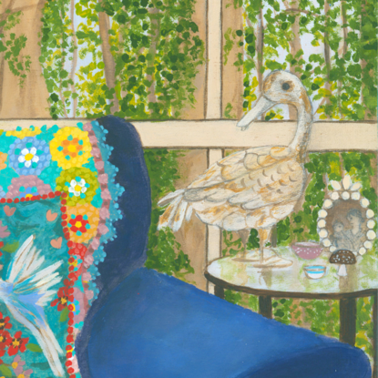 Interior scene with a rusty duck ornament on a glass side table, next to a blue arm chair and a rainbow crochet blanket hanging over the back behind a blue cushion featuring white birds and pink and red flowers. Against a window backdrop featuring leafy green trees