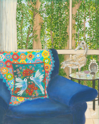 Interior scene with a rusty duck ornament on a glass side table, next to a blue arm chair and a rainbow crochet blanket hanging over the back behind a blue cushion featuring white birds and pink and red flowers. Against a window backdrop featuring leafy green trees