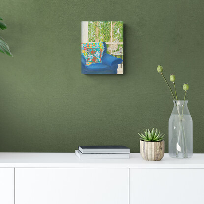 Interior scene with a rusty duck ornament on a glass side table, next to a blue arm chair and a rainbow crochet blanket hanging over the back behind a blue cushion featuring white birds and pink and red flowers. Against a window backdrop featuring leafy green trees