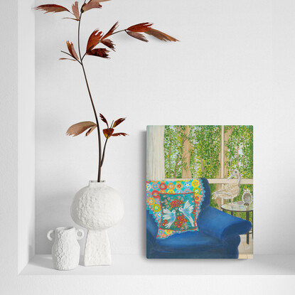 Interior scene with a rusty duck ornament on a glass side table, next to a blue arm chair and a rainbow crochet blanket hanging over the back behind a blue cushion featuring white birds and pink and red flowers. Against a window backdrop featuring leafy green trees