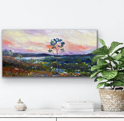 A distant vista of mountains and hills bathed in the evening light of the sunset and splashes of red earth against the greens of the grass and gum trees dotting the landscape. There is a distant river winding in the background. Framed view for illustration purposes only. 