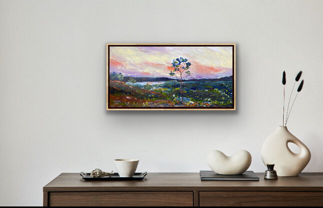 A distant vista of mountains and hills bathed in the evening light of the sunset and splashes of red earth against the greens of the grass and gum trees dotting the landscape. There is a distant river winding in the background. Framed view for illustration purposes only. 