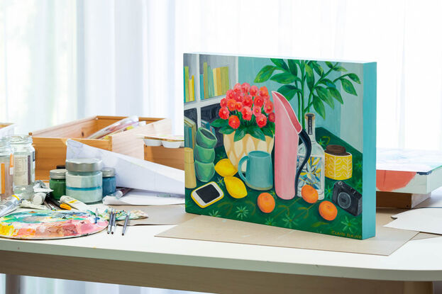 Table scene of pink Fink jug, gin bottle, potted plant, mug and lemons and mandarins. An i-phone sits on the table on the left on a green tablecloth and a camera and sugar bowl are on the right hand side. There is the suggestion of a bookshelf in the background.