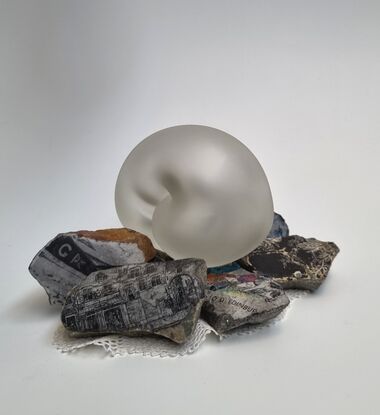 Rebecca has sculped an embryotic glass form to represent the innocence and fragility of life.
The glass form has been etched giving the glass a satin texture.
 She has incorporated rocks and stones, found objects such as images and fabric found in the family home. 
The embriotic form rests delicately on this mound of rubble exposed to the outside elements.
