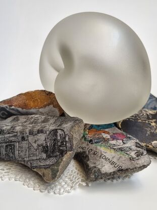 Rebecca has sculped an embryotic glass form to represent the innocence and fragility of life.
The glass form has been etched giving the glass a satin texture.
 She has incorporated rocks and stones, found objects such as images and fabric found in the family home. 
The embriotic form rests delicately on this mound of rubble exposed to the outside elements.
