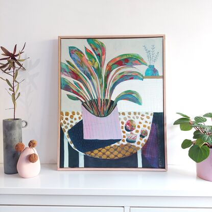 A light purple pot is filled with colourful foliage. Placed.on a spotty tablecloth and deep.shaodows beneath the table. Collage and pattern and markmaking are all part of this piece.