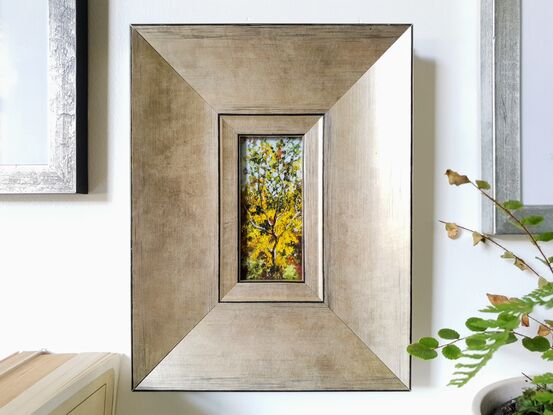 Framed miniature oil painting of a sunlit wattle sapling, surrounded by Australian bush in full bloom. Main colour yellow with green and pale blue. By Victoria Collins. Frame is wide edge, brushed gold with black detailing. 