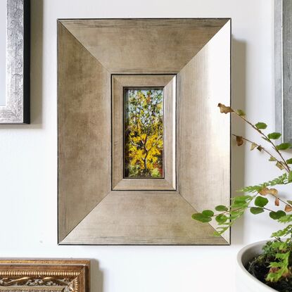 Framed miniature oil painting of a sunlit wattle sapling, surrounded by Australian bush in full bloom. Main colour yellow with green and pale blue. By Victoria Collins. Frame is wide edge, brushed gold with black detailing. 