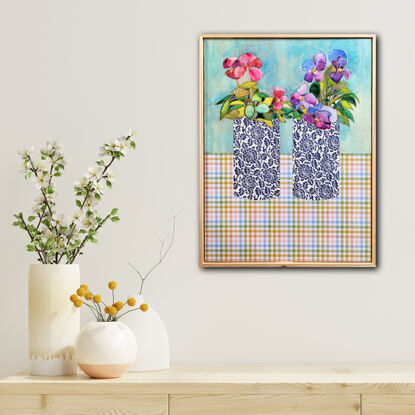 Two blue and white vases sit on a multicoloured checkered tablecloth. In the right vase sits purple flowers with gold centres, and the left are hot pink flowers with gold centres.