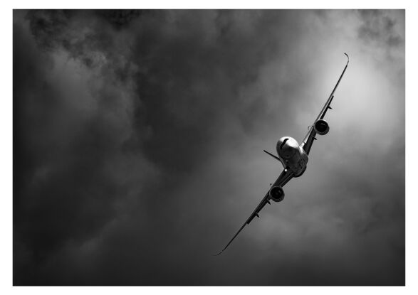 A black and white photograph of the Airbus A350, flying towards the photographer, in a steep right-hand turn in front of a dramatic cloudy background.