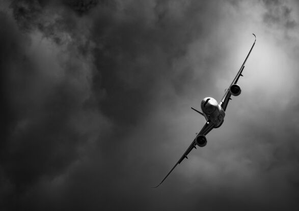 A black and white photograph of the Airbus A350, flying towards the photographer, in a steep right-hand turn in front of a dramatic cloudy background.