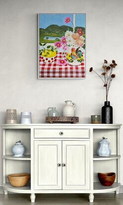 still life scene featuring a vase with a rooster filled with pink and white cosmos and orange roses next to a bowl with red spots filled with lemons, against a hinterland inspired landscape window view backdrop and a red gingham table cloth