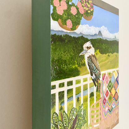 Kookaburra sitting on a verandah fence against a landscape backdrop inspired by the Sunshine Coast hinterland. Including plants and a cosy chair with colourful crochet blanket in the forecground.