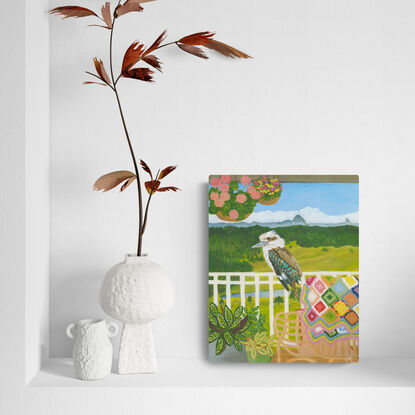 Kookaburra sitting on a verandah fence against a landscape backdrop inspired by the Sunshine Coast hinterland. Including plants and a cosy chair with colourful crochet blanket in the forecground.