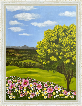 golden wattle tree and a field of pink yellow and white wild flowers against a landscape backdrop of rolling green hills and a blue sky