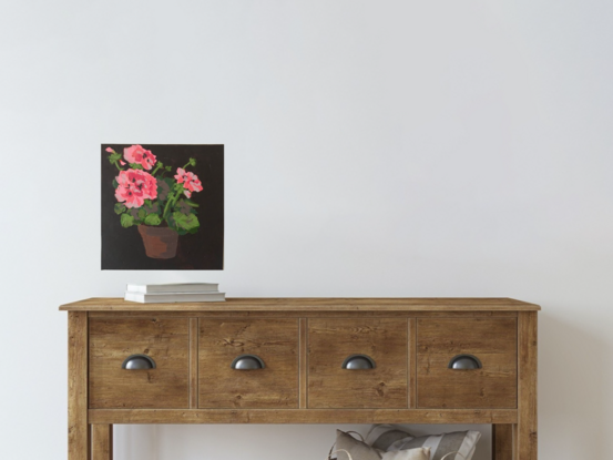 A terracotta pot sits in the centre of frame and in it is a pink geranium plant with big clusters of blooms sitting above scalloped edge mid green leaves, the background is a deep brown.black