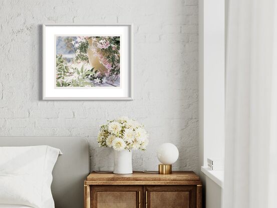 A vintage yellow ceramic jug is mostly in the right-hand half of the frame. It is side on with the lip facing inwards. It is outside but on a floor of old broken bathroom tiles. The background is out of focus but indicates a body of water and bush beyond. The jug is overflowing with jasmine. It spills out and falls all around. There is a lot of jasmine foliage with new bright buds in the middle and foreground of the left side of frame. The colours of the image are desaturated, except for the pinks of the jasmine stamen and the yellow jug.